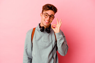 Young student man isolated on pink background winks an eye and holds an okay gesture with hand.