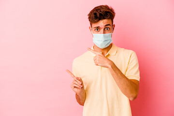 Young caucasian man wearing a protection for coronavirus isolated on pink background shocked pointing with index fingers to a copy space.