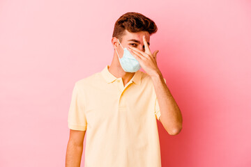 Young caucasian man wearing a protection for coronavirus isolated on pink background blink at the camera through fingers, embarrassed covering face.