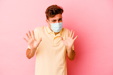 Young caucasian man wearing a protection for coronavirus isolated on pink background rejecting someone showing a gesture of disgust.