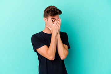 Young caucasian man isolated on blue background blink through fingers frightened and nervous.