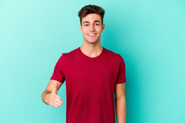 Young caucasian man isolated on blue background stretching hand at camera in greeting gesture.