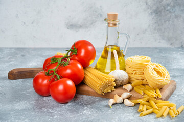 Assortment of raw pasta, olive oil and tomatoes on wooden board
