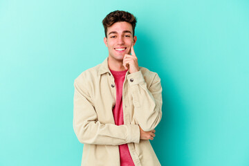 Young caucasian man isolated on blue background smiling happy and confident, touching chin with...