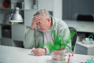 At the end of the working day, an elderly office worker felt a headache, he sits at his desk and holds his head.