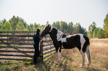 In autumn, a girl ties a horse to a fence on a sunny day.
