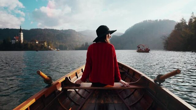 Summer vacation ispiration. Romantic woman in hat sitting in boat on Bled lake at hazy scenic sunset. Rear view of a girl relaxing while travel adventure. Freedom, experience, sightseeing concept.