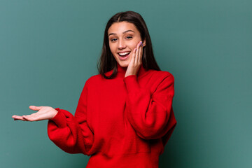 Young Indian woman isolated on blue background holds copy space on a palm, keep hand over cheek....