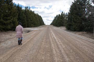 Obraz premium A woman wearing a pink puff parka and a black toque and jeans walks down an old dirt road in the countryside lined with coniferous spruce trees in March, 2021, Ontario Canada.