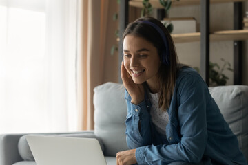 Close up smiling woman wearing headphones chatting, using laptop, looking at computer screen, sitting on couch at home, happy young female making video call to relatives or friends, talking