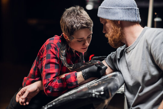 In The Interior Of A Tattoo Parlor, A Young Man With A Beard Is Tattooed On His Arm Using The Stick And Poke Method. Young Artist Girl In A Checkered Shirt