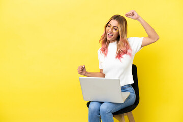 Young woman sitting on a chair with laptop over isolated yellow background celebrating a victory