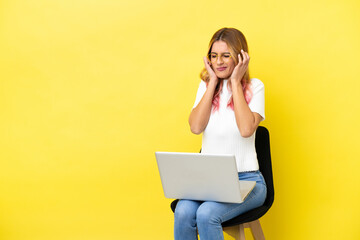 Young woman sitting on a chair with laptop over isolated yellow background frustrated and covering ears