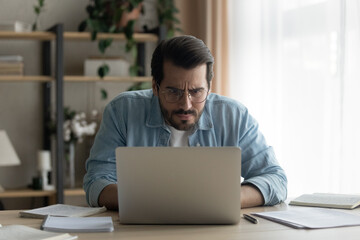 Close up puzzled focused businessman wearing glasses looking at laptop screen, employee or freelancer working on difficult research project online, reading bad news, analyzing financial report