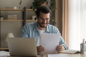 Close up smiling businessman wearing glasses reading good news in paper letter, sitting at work desk with laptop, happy satisfied man holding documents, notification, job promotion, loan approval