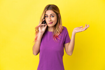Young woman using mobile phone over isolated yellow background having doubts while raising hands