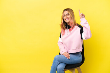 Young woman sitting on a chair over isolated yellow background showing and lifting a finger in sign of the best