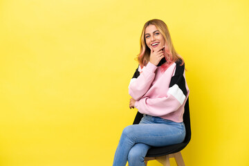 Young woman sitting on a chair over isolated yellow background happy and smiling