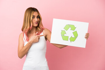 Young woman over isolated pink background holding a placard with recycle icon and  pointing it