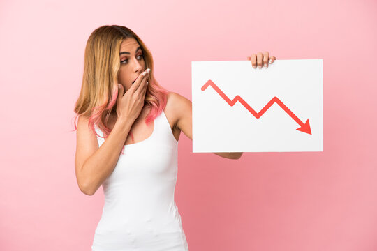 Young woman over isolated pink background holding a sign with a decreasing statistics arrow symbol with surprised expression