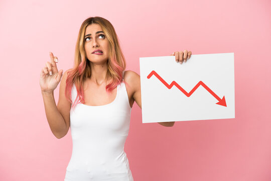 Young woman over isolated pink background holding a sign with a decreasing statistics arrow symbol with fingers crossing