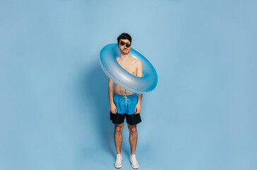 Full length photo of a foolish funny caucasian guy in shorts and without a t-shirt with a inflatable blue ring and crooked glasses stands on a blue isolated background, silly mood, joke