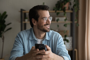 Close up smiling dreamy man wearing glasses distracted from phone, looking to aside, holding smartphone, visualizing good future, waiting for call or message, enjoying leisure time with gadget