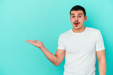 Young caucasian man isolated on blue background impressed holding copy space on palm.