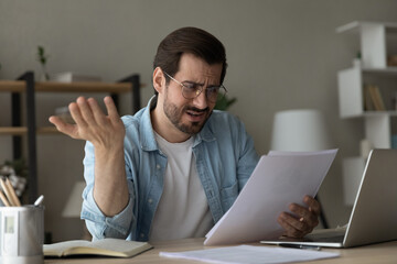 Close up unhappy dissatisfied man wearing glasses reading bad news, holding letter, checking financial documents, sitting at work table, stressed businessman worried about money problem or debt