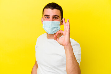 Young caucasian man wearing a protection for coronavirus isolated on yellow background cheerful and confident showing ok gesture.
