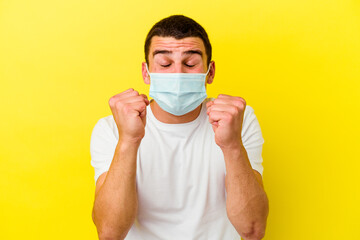 Young caucasian man wearing a protection for coronavirus isolated on yellow background raising fist, feeling happy and successful. Victory concept.