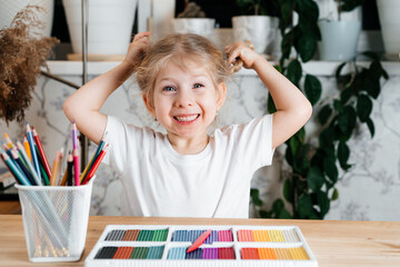 a small blonde girl with a charming smile sits at a table with a bright colorful palette of colored plasticine for modeling at home, hobbies and child development