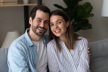 Head shot portrait smiling couple family hugging, looking at camera, sitting on couch at home, happy woman and man making video call to relatives or friends, positive bloggers shooting vlog