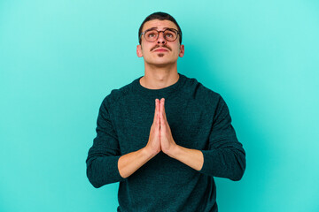 Young caucasian man isolated on blue background holding hands in pray near mouth, feels confident.