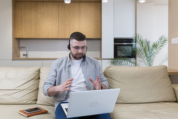 A man with a beard works from home sitting on the couch, uses a laptop and a headset with a microphone conducts an online consultation