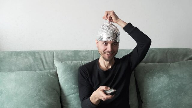 Conspiracy theorist man wearing tin foil hat watching TV on couch at home. Strange guy belief that aluminium cap shield brain from mind control, electromagnetic fields of fake news.