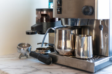 An espresso machine sits on a white and grey restaurant counter. The coffee machine or aluminum appliance is used to manufacture hot cafe coffee. There are two jugs and a coffee press on the maker.