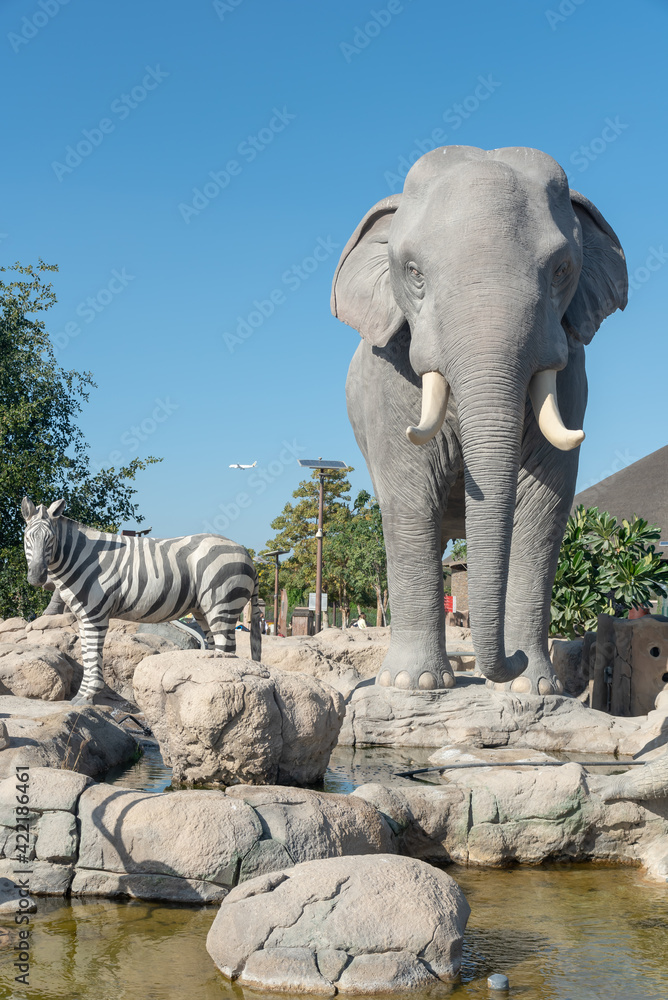 Wall mural Vertical shot of statues of a big elephant and zebra near the water under the blue sky in Dubai zoo