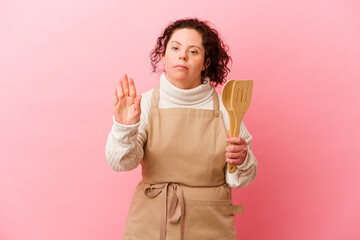 Woman with Down syndrome cooking at home isolated on pink background standing with outstretched hand showing stop sign, preventing you.