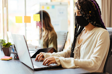 Business women wearing protective masks sitting at their desks separated by plexiglass dividers