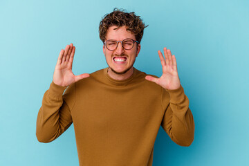 Young caucasian man wearing eyeglasses isolated on blue background screaming with rage.
