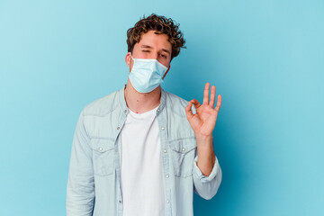 Young caucasian man wearing an antiviral mask isolated on blue background winks an eye and holds an okay gesture with hand.