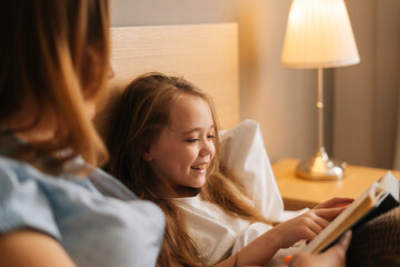 Close-up of mother and cheerful adorable daughter reading together children book before going to...