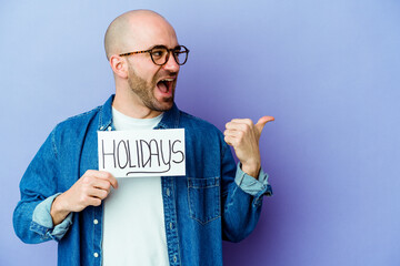 Young caucasian bald man holding a holidays placard isolated on blue background points with thumb finger away, laughing and carefree.