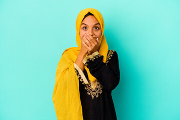Young muslim woman isolated on blue background shocked covering mouth with hands.