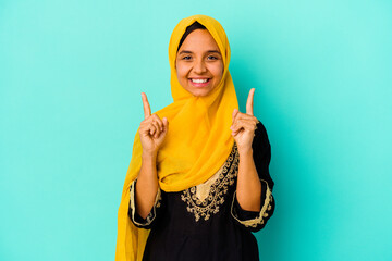 Young muslim woman isolated on blue background indicates with both fore fingers up showing a blank...