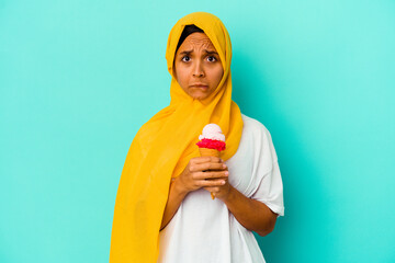 Young muslim woman eating an ice cream isolated on blue background confused, feels doubtful and...
