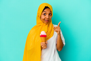 Young muslim woman eating an ice cream isolated on blue background having an idea, inspiration concept.