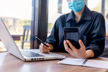 Young woman with protective face mask sitting in coffee shop at wood table using smartphone.and laptop. Girl browsing internet, chatting, blogging. Female holding phone and looking on screen