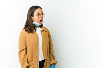 Young latin woman wearing a mask to protect from covid isolated on white background being shocked because of something she has seen.